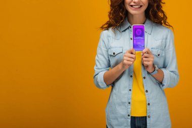 turuncu ekranda e alışveriş uygulaması ile akıllı telefon tutan neşeli kıvırcık kadın kırpılmış görünümü 