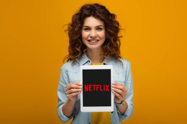 turuncu üzerinde dururken ekranda netflix uygulaması ile dijital tablet tutan neşeli kızıl saçlı kadın 