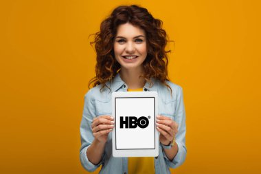 turuncu üzerinde dururken ekranda hbo uygulaması ile dijital tablet tutan mutlu kızıl saçlı kadın 
