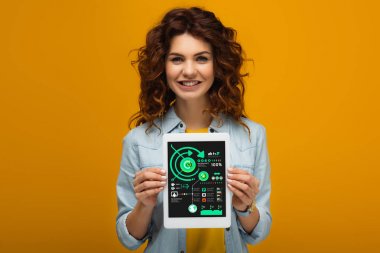 turuncu üzerinde dururken ekranda grafikler ve grafikler ile dijital tablet tutan gülümseyen kızıl saçlı kadın 
