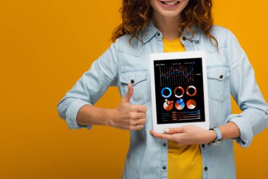 ekranda grafikler ve grafikler ile dijital tablet tutan ve turuncu izole başparmak gösteren neşeli kıvırcık kız kırpılmış görünümü 