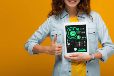 ekranda grafikler ve grafikler ile dijital tablet tutan ve turuncu izole başparmak gösteren neşeli kıvırcık kadın kırpılmış görünümü 
