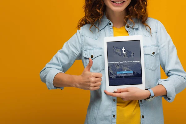 裁剪视图欢快的卷发女孩拿着数字平板电脑与Tumblr应用程序在屏幕上 并显示大拇指隔离在橙色 — 图库照片