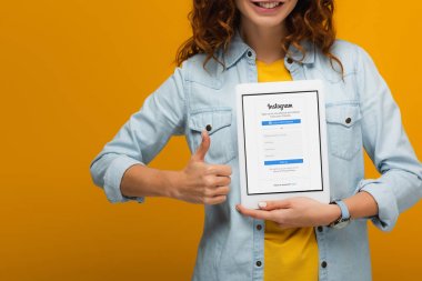 ekranda instagram uygulaması ile dijital tablet tutan ve turuncu izole başparmak gösteren neşeli kıvırcık kadın kırpılmış görünümü 