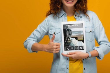 ekranda bilet uygulaması ile dijital tablet tutan ve turuncu izole başparmak gösteren neşeli kıvırcık kadın kırpılmış görünümü 