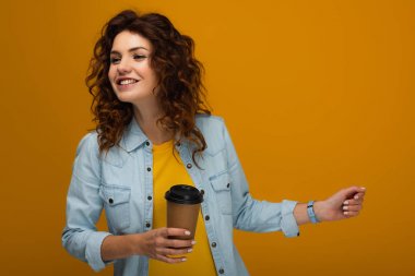 happy curly redhead girl holding coffee to go isolated on orange  clipart