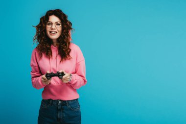 happy curly redhead girl playing video game while holding joystick on blue clipart
