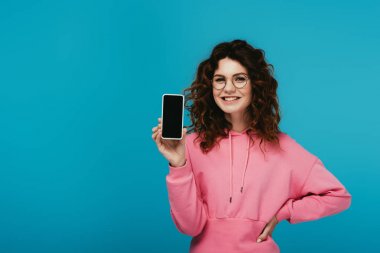 gözlük neşeli kıvırcık kız kalça eliyle ayakta ve mavi boş ekran ile akıllı telefon tutarak 