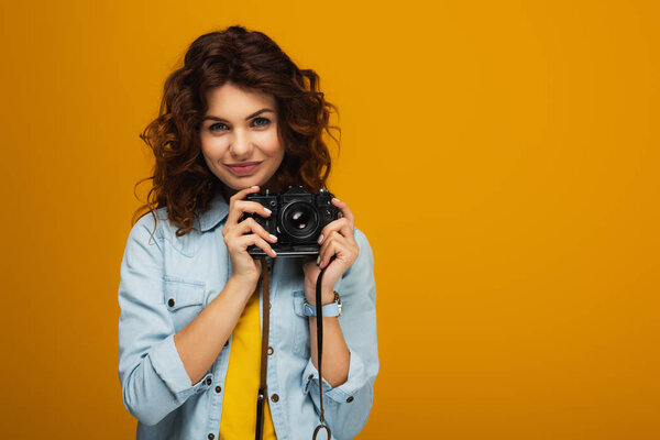 curly redhead photographer holding digital camera isolated on orange 