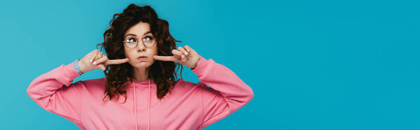 панорамный снимок кудрявой рыжей девушки в очках, держащей пальцы возле щек, изолированных на голубом
 