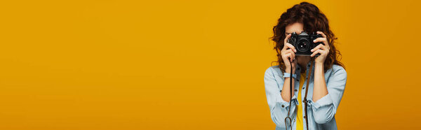 panoramic shot of curly redhead photographer covering face with digital camera isolated on orange 