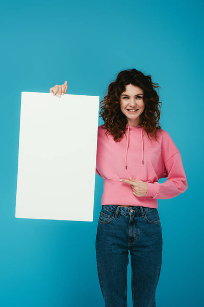 счастливая кудрявая рыжая женщина, указывающая пальцем на пустой плакат на синем
 