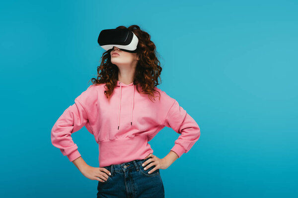 curly girl wearing virtual reality headset while standing with hands on hips on blue 
