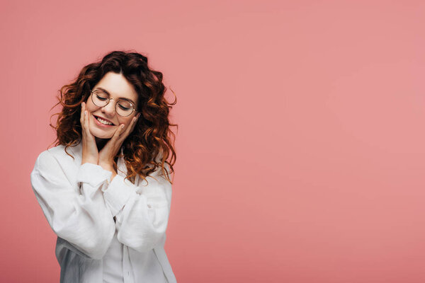 happy curly woman in glasses smiling while touching face on pink