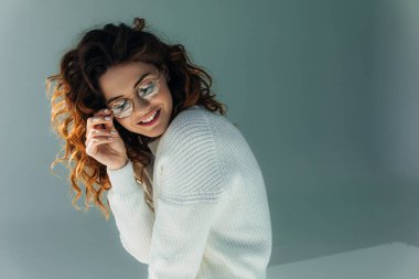 cheerful curly redhead girl touching glasses and smiling on grey  clipart