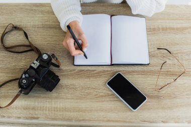 boş ekranlı boş dizüstü bilgisayarın, dijital kameranın, gözlüklerin ve akıllı telefonun yanında kalem tutan genç kadının kırpılmış görünümü 