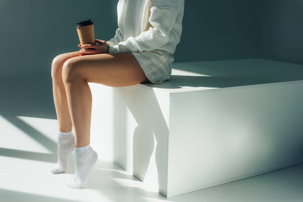 cropped view of young woman sitting in socks and holding paper cup on grey