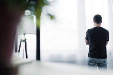 selective focus of short haired man in black t-shirt standing by window at home clipart