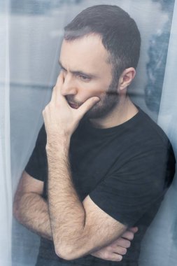 handsome, pensive man in black t-shirt standing by window and holding hand on mouth clipart