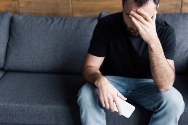 depressed man sitting on sofa with smartphone and holding hand on forehead