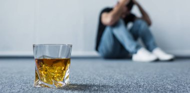 selective focus of depressed man sitting on floor by wall near glass of whiskey clipart