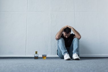 depressed man sitting on floor by white wall near bottle and glass of whiskey clipart
