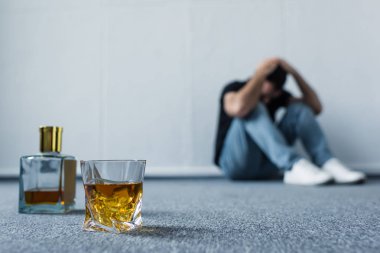 selective focus of suffering man sitting on floor by white wall near bottle and glass with whiskey clipart