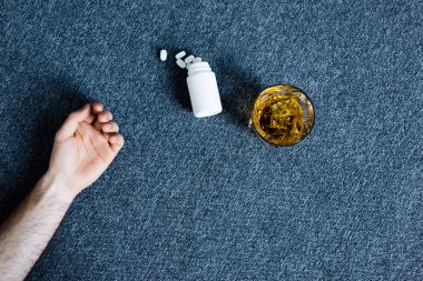 cropped view of man lying on grey floor near glass of whiskey and container with pills