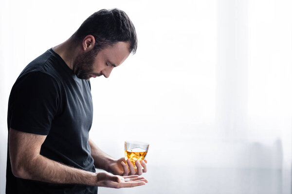 depressed man in black t-shirt standing with glass of whiskey and handful of pills