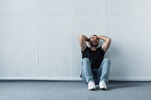 adult depressed man screaming while sitting on floor by white wall 