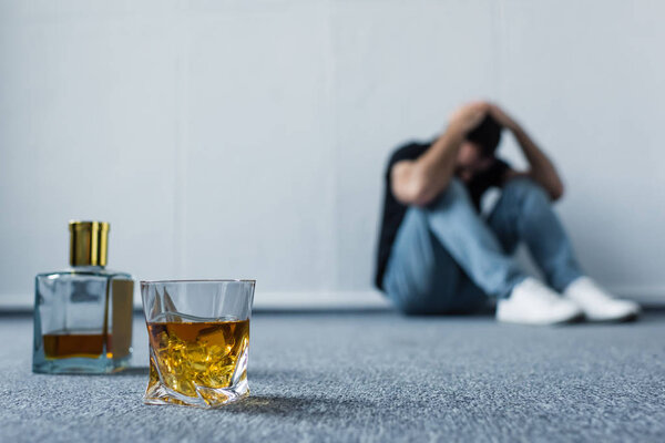 selective focus of suffering man sitting on floor by white wall near bottle and glass with whiskey