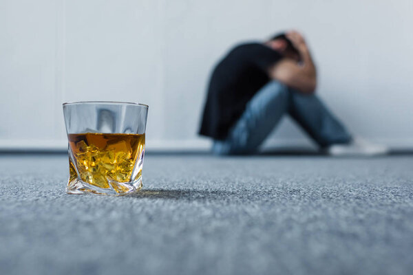 селективный фокус депрессивного мужчины, страдающего, сидя на сером полу возле стакана виски
