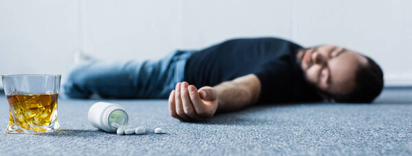 panoramic shot of unconscious man lying on grey floor near glass of whiskey and container with pills 