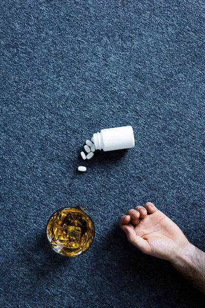 top view of male hand near glass of whiskey and container with pills on grey floor