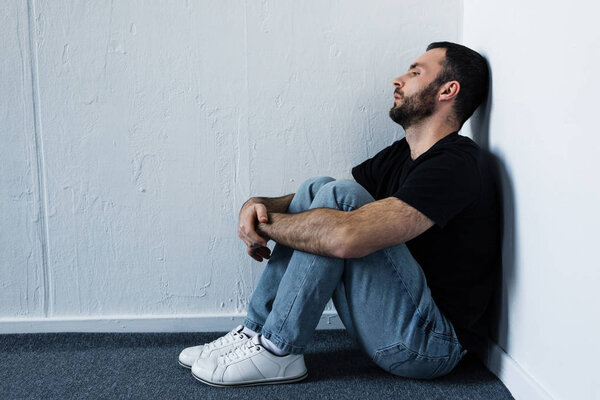 handsome depressed man sitting on floor in corner by white wall and looking away