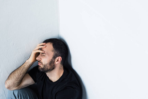 depressed man sitting in corner with closed eyes and holding hand on forehead