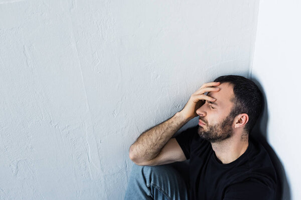 high angle view of upset man sitting in corner with closed eyes and holding hand on forehead