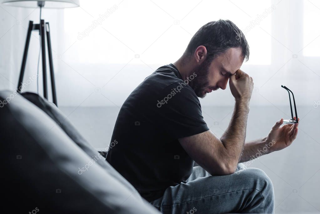 upset man sitting on sofa with closed eyes and holding glasses