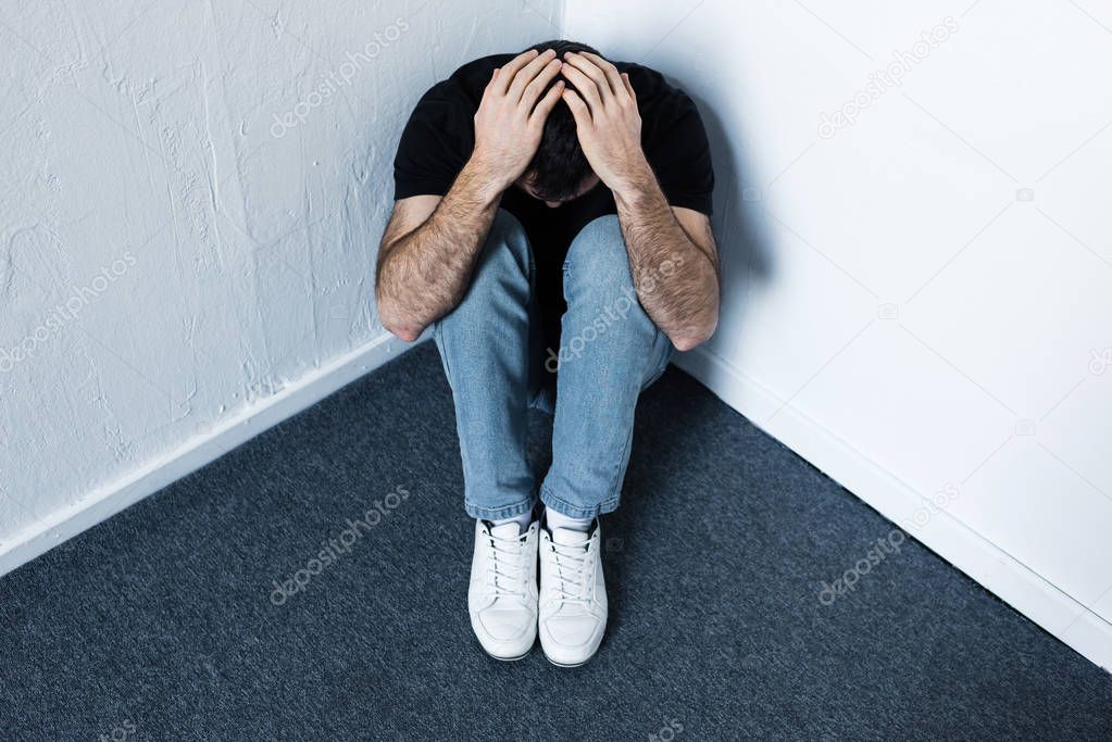 depressed man sitting on grey floor in corner and holding hands on head