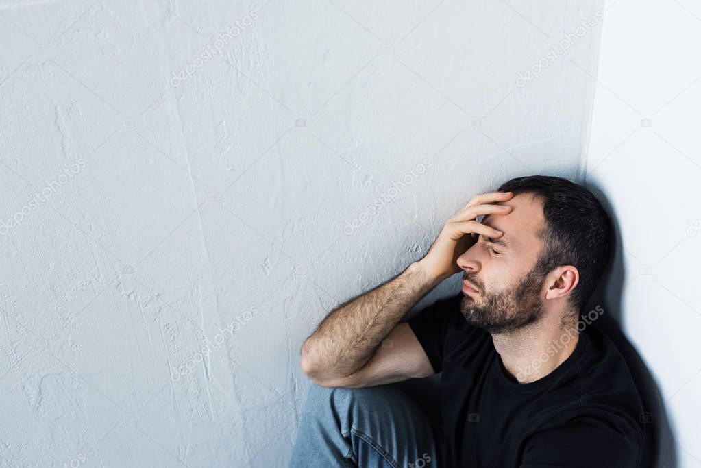 high angle view of upset man sitting in corner with closed eyes and holding hand on forehead