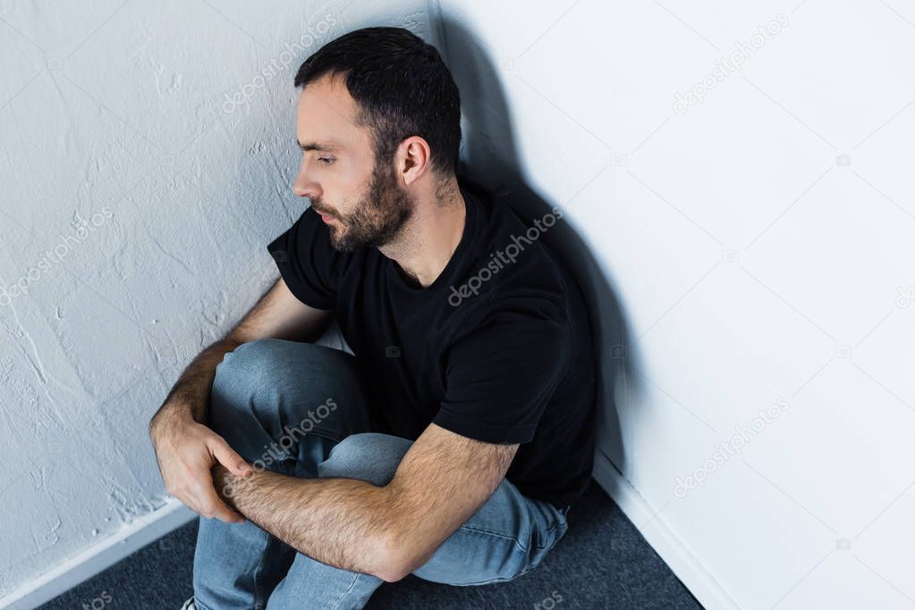 handsome bearded depressed man sitting on floor in corner and looking away