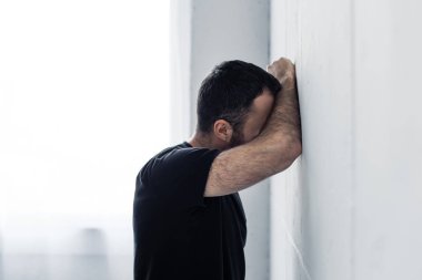adult depressed man in black t-shirts standing near white wall at home clipart