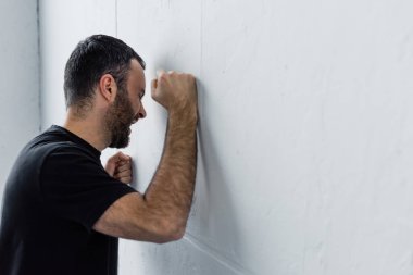 adult depressed man screaming while standing by white wall at home clipart