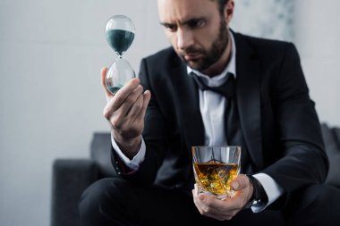 viski bardağı tutarken kum saatibakarak takım elbise üzgün sakallı adam seçici odak