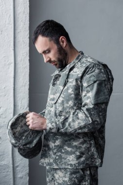 sad bearded man in military uniform standing with bowed had and holding cap in hands clipart
