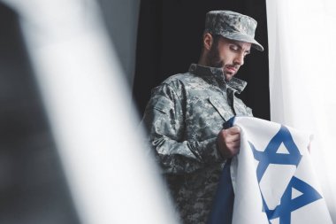 selective focus of thoughtful military man in uniform holding israel national flag while standing by window clipart