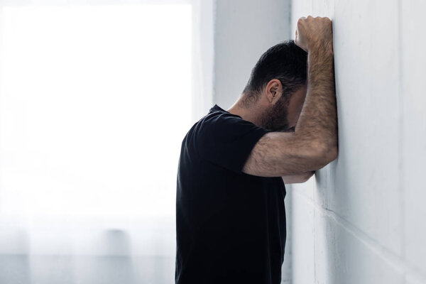 adult depressed man in black t-shirt suffering while standing near white wall