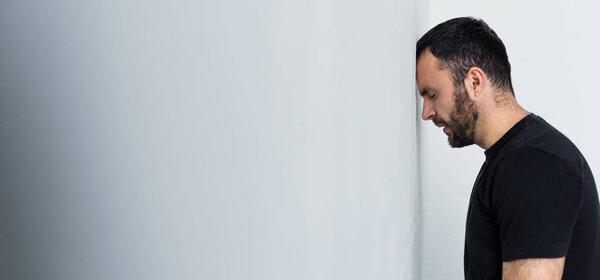 panoramic shot of depressed bearded man standing by white wall with closed eyes