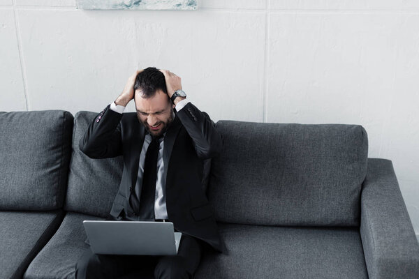 сердитый бизнесмен, держась за голову, сидя на диване и используя смартфон
