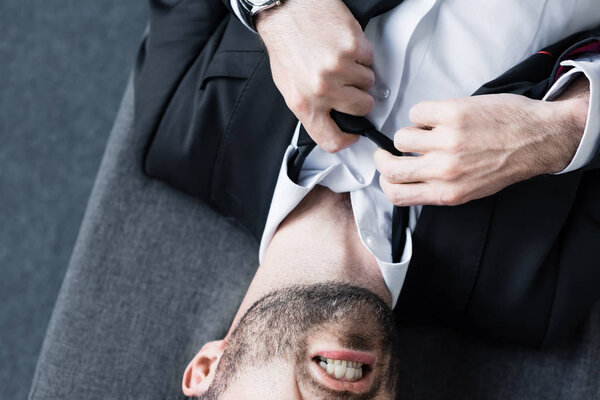 partial view of depressed businessman suffering and untying tie while lying on sofa